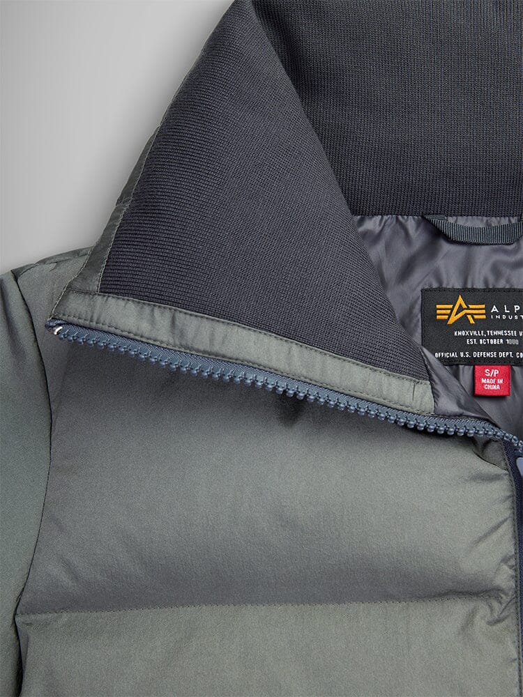 A close-up of a dark green Sierra Short Puffer W parka with a black ribbed collar highlights its water-resistant material. Inside, a label features an emblem with the words "Alpha Industries" in yellow and white text.