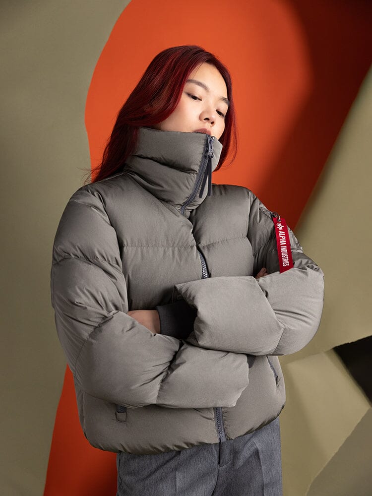 An individual with long hair dons the gray, water-resistant Sierra Short Puffer W by Alpha Industries, standing with their arms crossed.