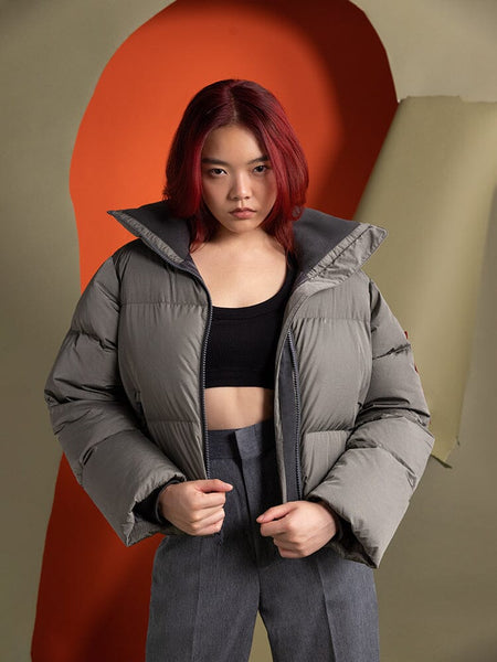 A person with red hair is wearing the Sierra Short Puffer W, a water-resistant gray jacket from Alpha Industries, over a black top.