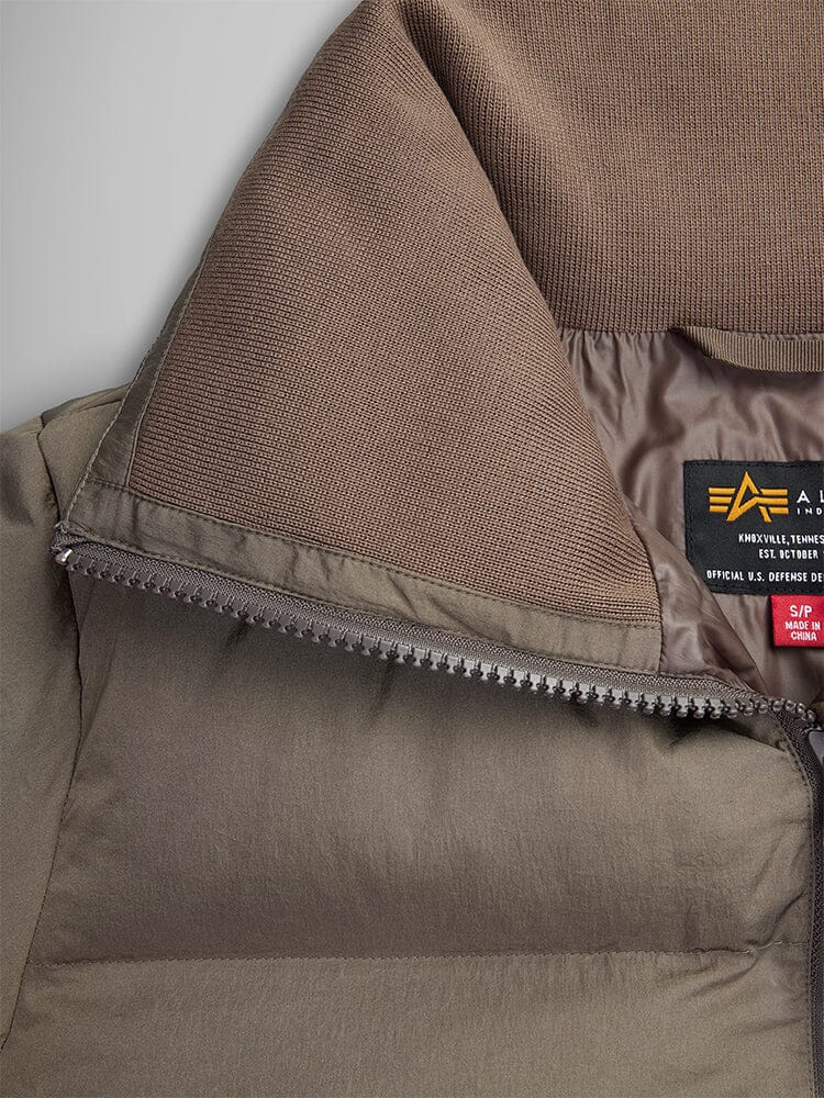 Close-up of a brown Sierra Short Puffer W from Alpha Industries, featuring a visible label and partially open zipper that reveals the down fill inner lining. The jacket's water-resistant fabric offers both style and protection from the elements.