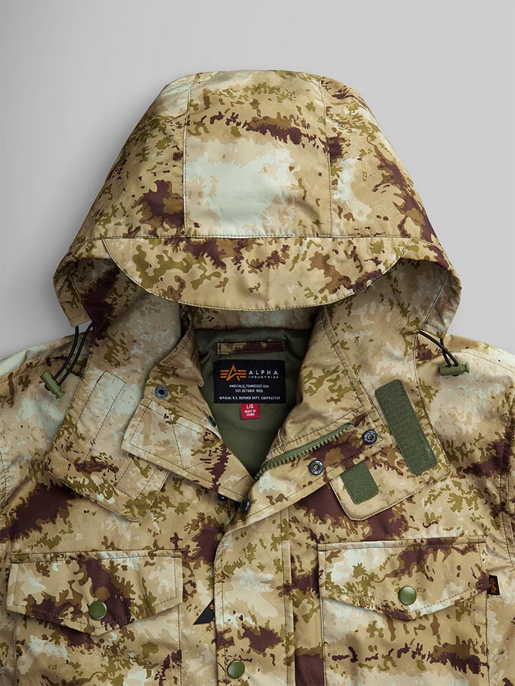 Close-up view of the Tactical M-65 Field Jacket in beige and brown camouflage from Alpha Industries. It includes multiple pockets and a black label on the inside collar, highlighting Alpha Industries' signature design.