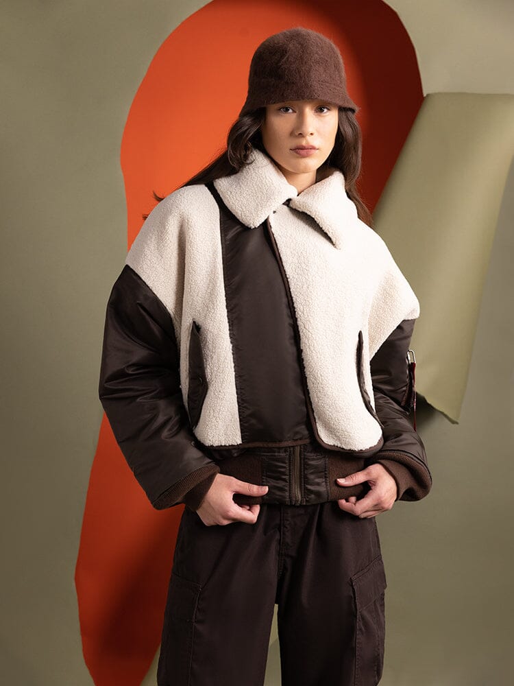 A person dressed in the Women's B-15 Sherpa Hybrid Bomber Jacket by Alpha Industries, featuring a brown and white color scheme and an asymmetrical zipper, paired with a brown hat.