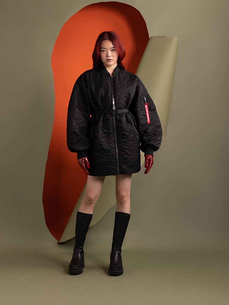 A person wearing the Alpha Industries Women's Camo Quilted Belted MA-1 Bomber Jacket in black stands, accompanied by red gloves and black boots.