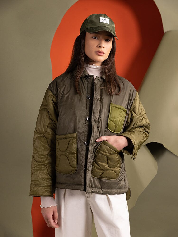 An individual wearing the Women's Insulated Chore Coat in OG-107 green from Alpha Industries, paired with light pants and a green cap.