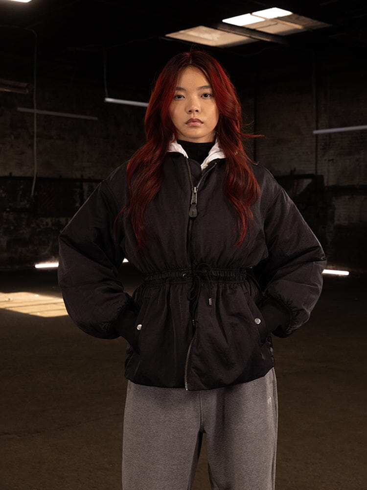 An individual with long red hair in the Women's Short Cinched Waist Jacket by Alpha Industries stands confidently in gray pants. The dimly lit room highlights the jacket's dark padded design, featuring a welt pocket and quilted lining, while the adjustable drawcord waist adds both style and functionality to their look.