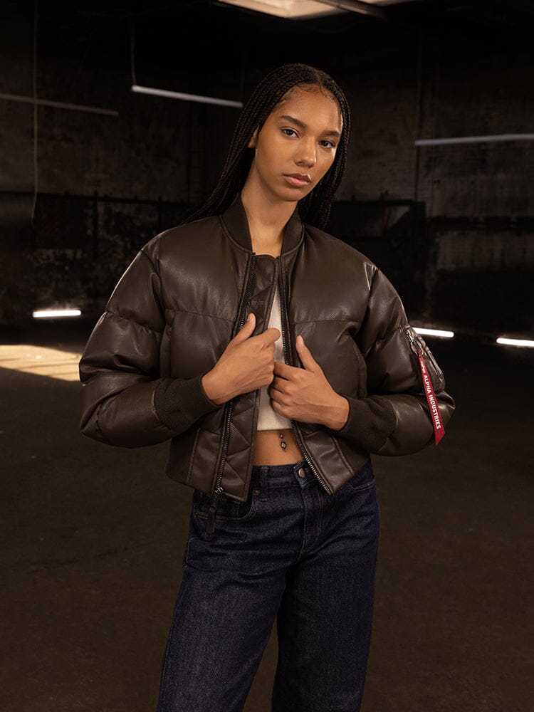 In the dimly lit room, a person stands confidently, hands gripping the open Women's Sierra Faux Leather Short Puffer Jacket. The brown bomber jacket from Alpha Industries pairs seamlessly with jeans, exuding a stylish charm.