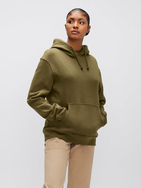 An individual wearing an Alpha Industries, Inc. Alpha Hoodie in unisex olive green, complete with an Alpha applique, and paired with beige pants to highlight the comfort of cotton fabric.