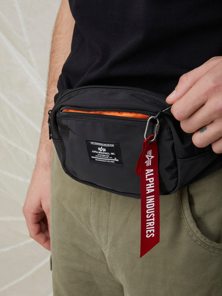 A person wearing the Alpha Industries CREW WAIST BAG from the FW24-DROP1 collection with an adjustable waist strap, over olive green pants and a black shirt.