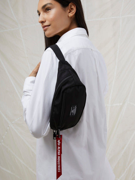 A person in a white shirt stands against a light background, wearing the sleek and versatile CREW WAIST BAG by Alpha Industries from their FW24-DROP1 collection. The bag features a distinctive red Alpha Industries tag and is crafted from flight nylon material, complete with an adjustable waist strap for diverse styling options.