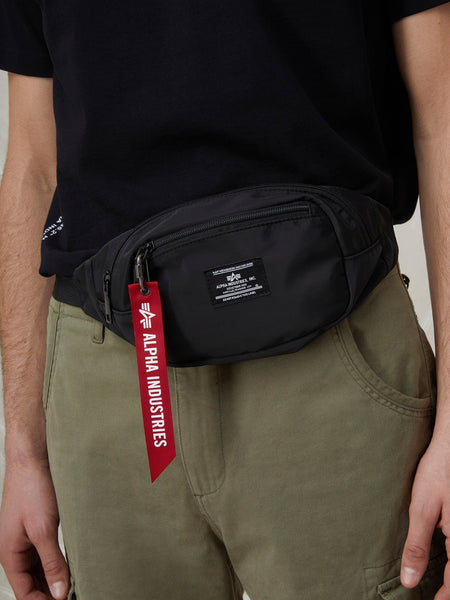 A person wearing an Alpha Industries CREW WAIST BAG from the FW24-DROP1 collection, featuring a sleek black design with the iconic red "Remove Before Flight" tag, is dressed in a black shirt and olive pants. The bag's flight nylon material and adjustable waist strap provide both style and functionality.