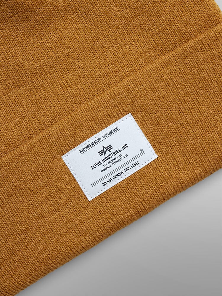 A mustard-colored knit Essential Beanie showcasing a white Alpha Industries, Inc. paper label stitched on one side.