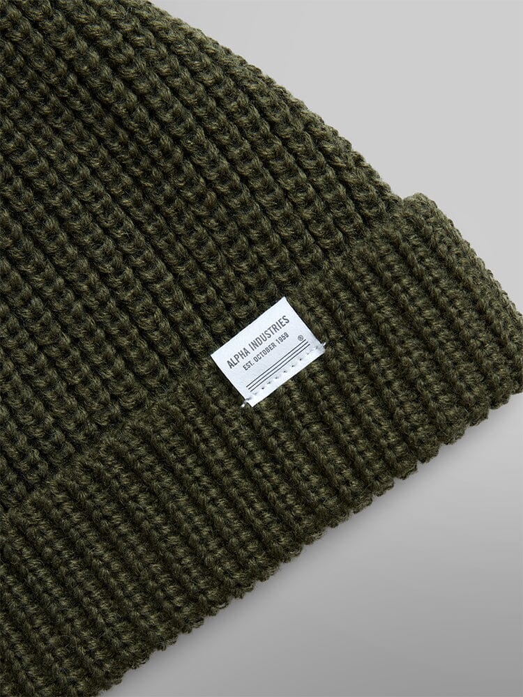 Close-up of the Essential Watch Cap in a green knit wool-blend with a folded brim, featuring a white kabel that says Alpha Industries.