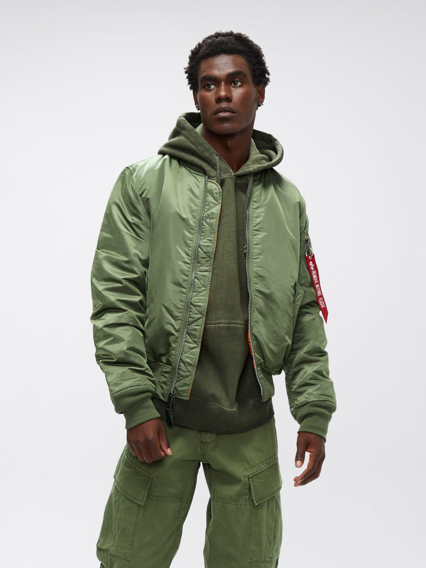 An individual wearing an Alpha Industries MA-1 Bomber Jacket Slim Fit in green, paired with a green hoodie and green cargo pants, gazes slightly to the side.