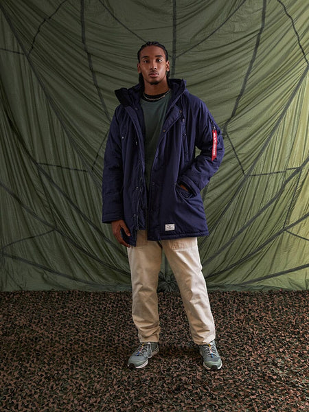 A person stands wearing the N-3B Altitude Mod Parka in blue from Alpha Industries over a green shirt.