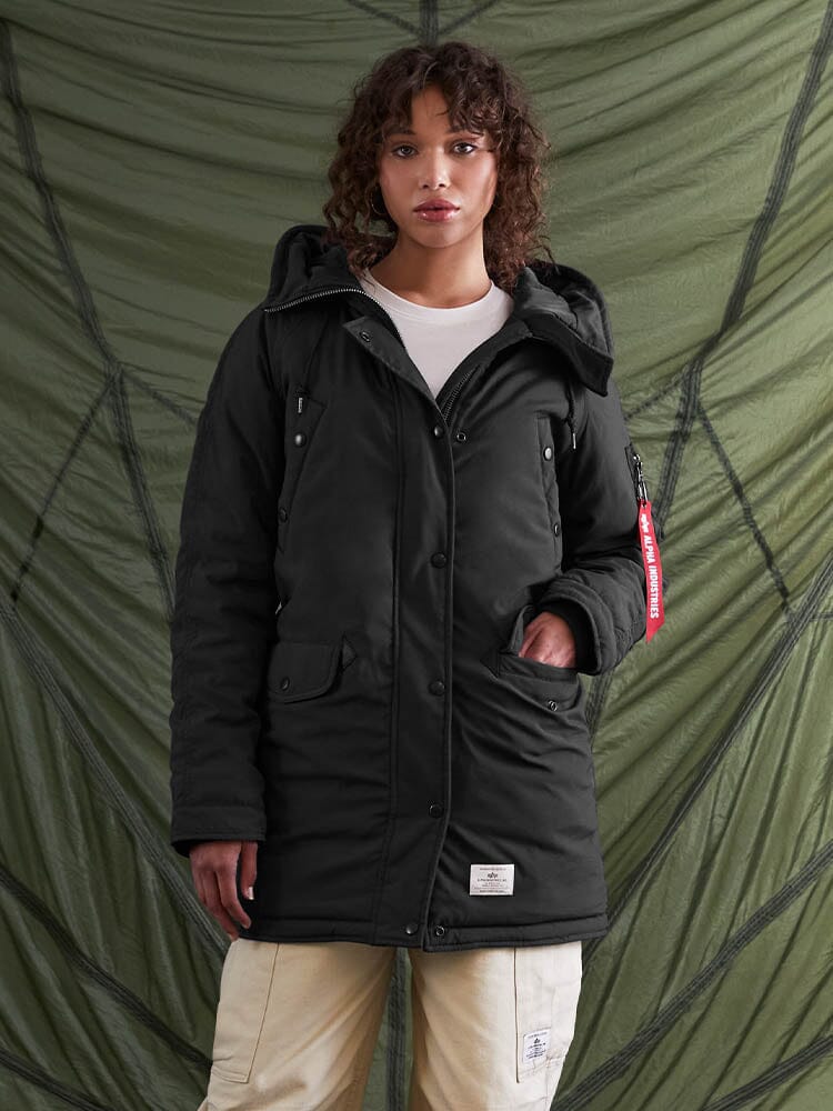 A person wearing the N-3B Altitude Mod Parka W by Alpha Industries, Inc., a US Navy-inspired black parka with a snorkel hood, paired with light-colored pants.