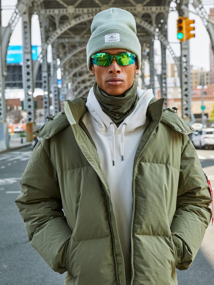 A person in an Alpha Industries Puffer Parka paired with reflective sunglasses stands under an urban bridge.