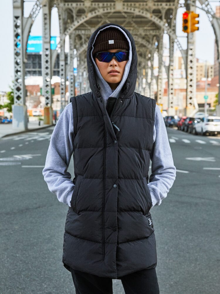 An individual clad in an Alpha Industries Sierra Convertible Jacket W, along with a gray hoodie, beanie, and sunglasses, stands on an urban street beneath a metal bridge.