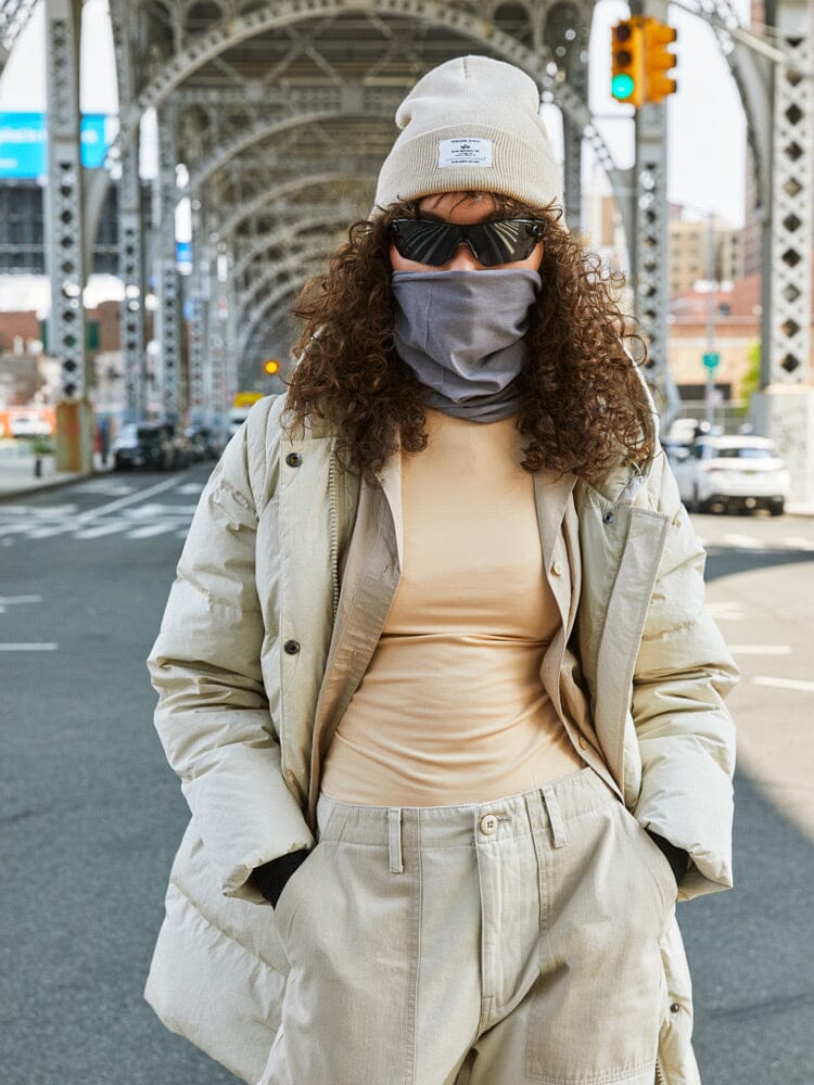 A person wearing an Alpha Industries Sierra Convertible Jacket W in beige and sunglasses stands on a city street under a metal bridge.