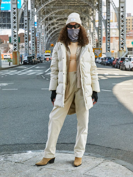 A person wearing the Sierra Convertible Jacket W in beige from Alpha Industries confidently stands under a bridge, their beanie and face covering completing the look.